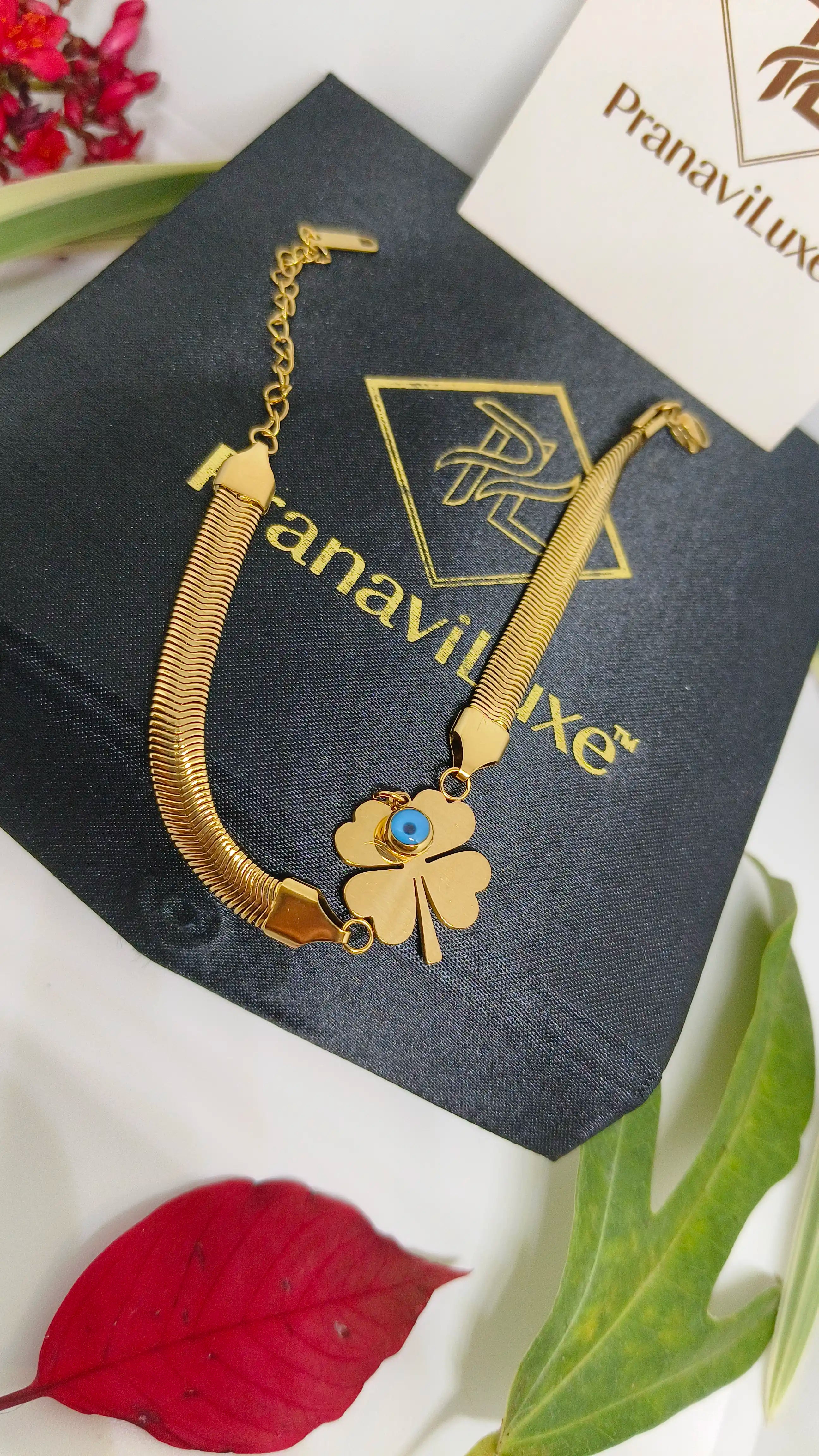 Evil's Eye Leaf Charm Snake Chain Bracelet in gold tone, featuring a four-leaf clover charm with an evil eye detail, displayed on a black branded box with floral accents.