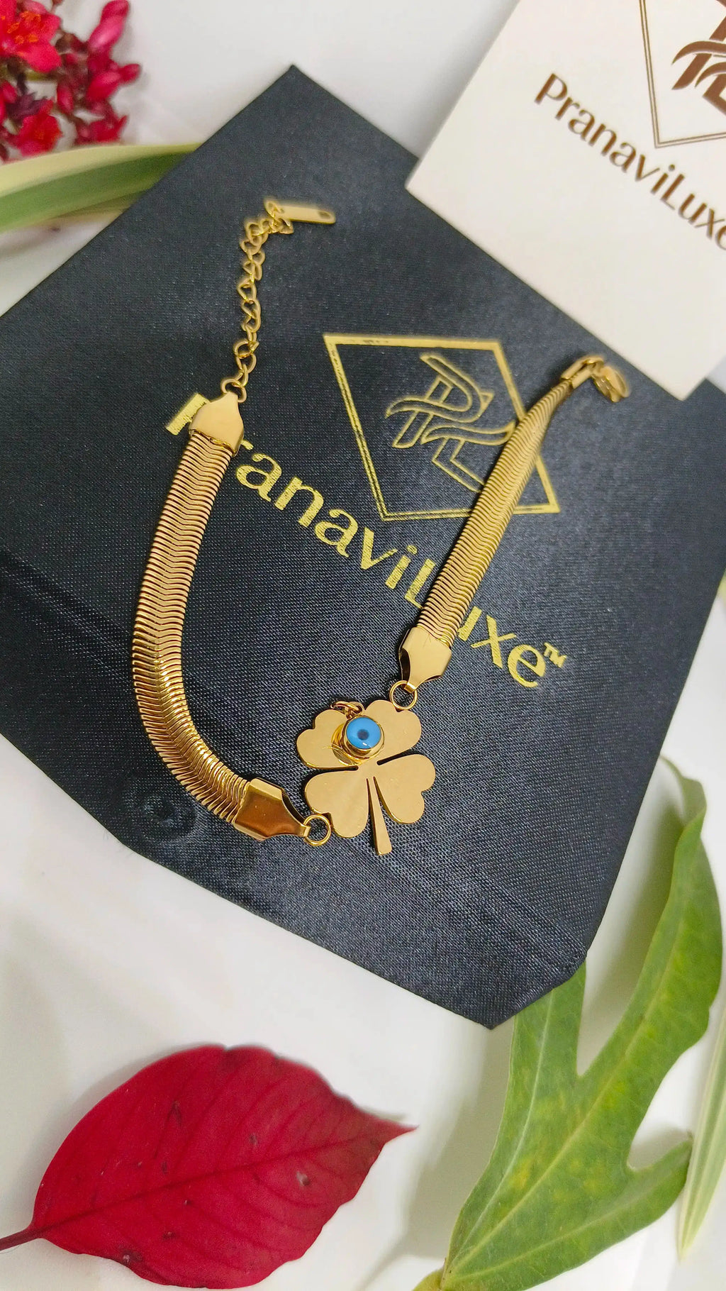 Evil's Eye Leaf Charm Snake Chain Bracelet in gold tone, featuring a four-leaf clover charm with an evil eye detail, displayed on a black branded box with floral accents.
