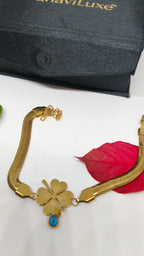 Elegant gold-toned Evil's Eye Leaf Charm Snake Chain Bracelet with a four-leaf clover pendant and blue evil eye charm, displayed on a white surface with a red leaf nearby and branded black packaging.