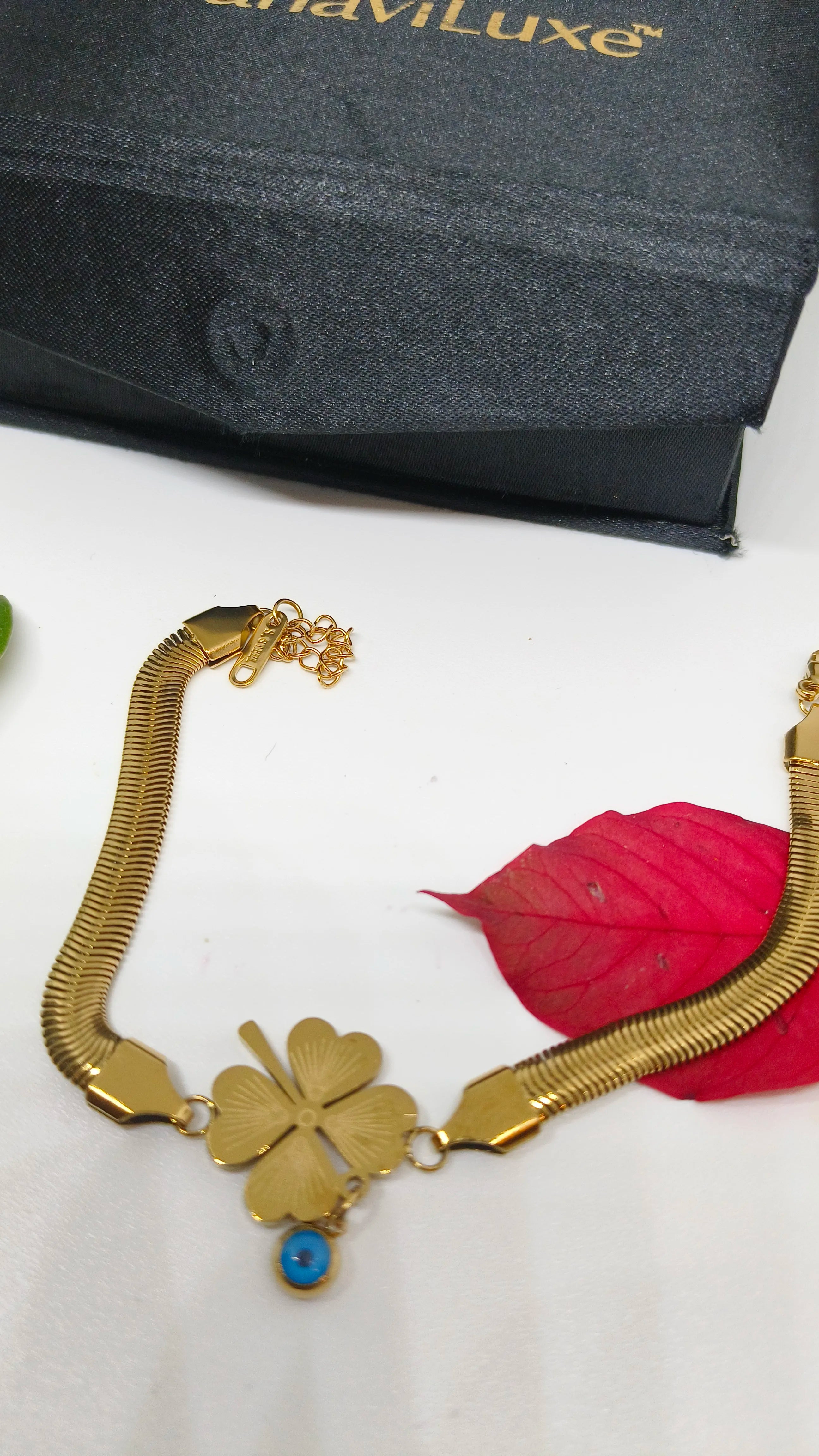 Elegant gold-toned Evil's Eye Leaf Charm Snake Chain Bracelet with a four-leaf clover pendant and blue evil eye charm, displayed on a white surface with a red leaf nearby and branded black packaging.