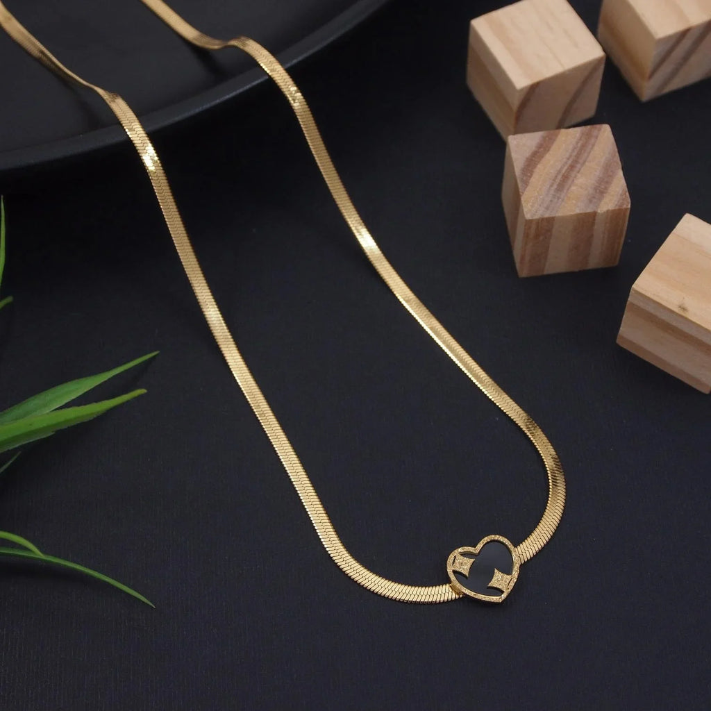 Gold necklace with a heart-shaped pendant on a dark surface with wooden blocks and green leaves.