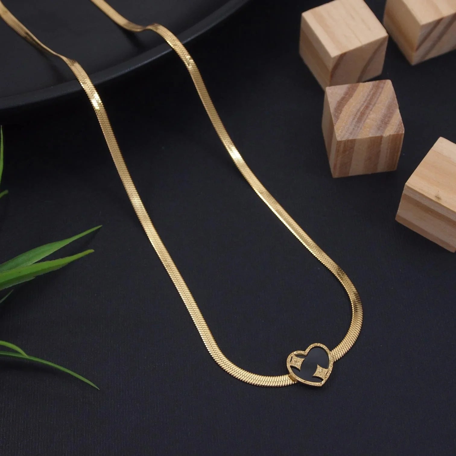 Gold necklace with a heart-shaped pendant on a dark surface with wooden blocks and green leaves.