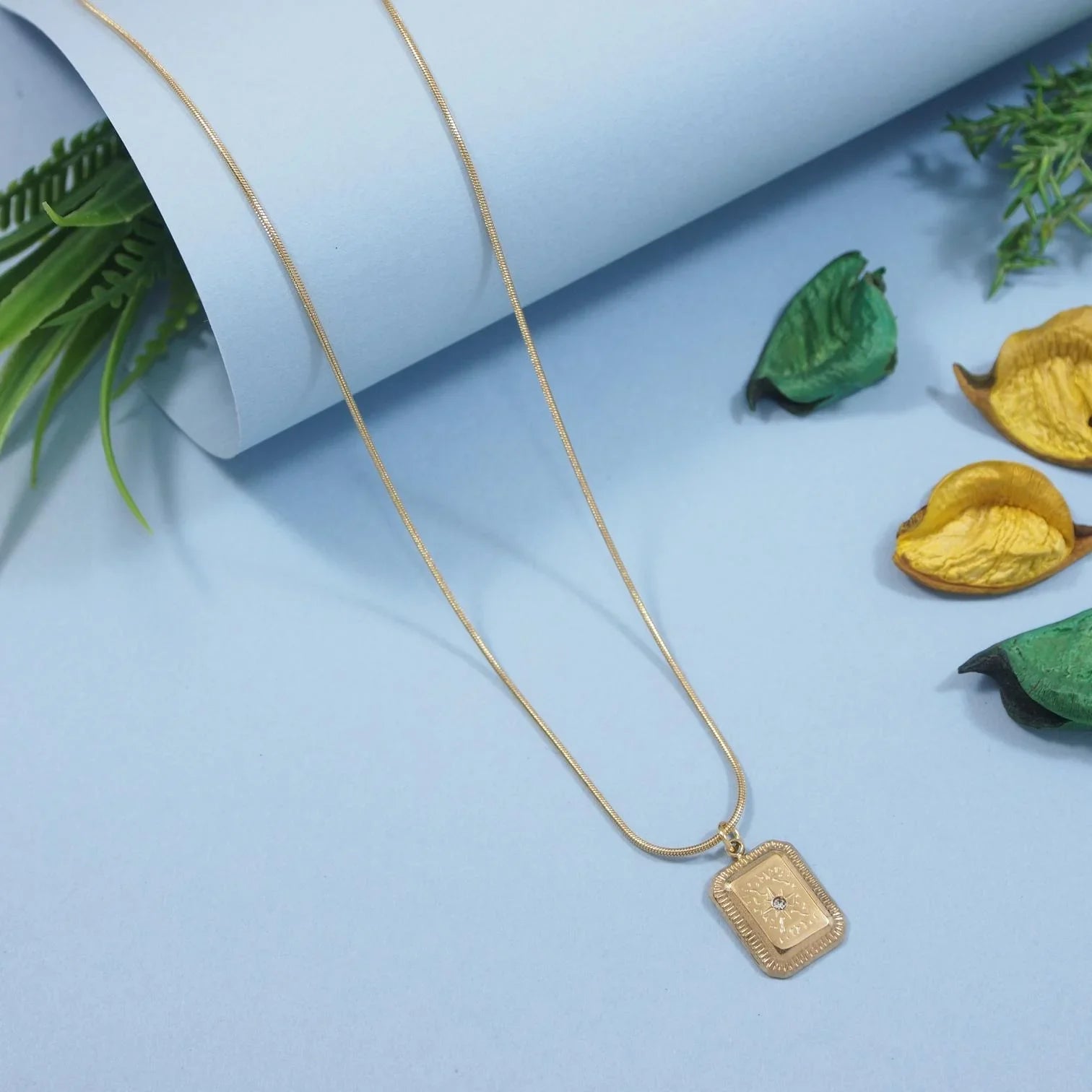 Gold necklace with a rectangular pendant on a light blue background with decorative leaves.