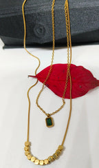 Green Stone Layered Necklace featuring a delicate gold chain with a small rectangular green stone pendant and a second gold chain with circular gold accents, displayed on a white surface with a red leaf and a black fabric background.