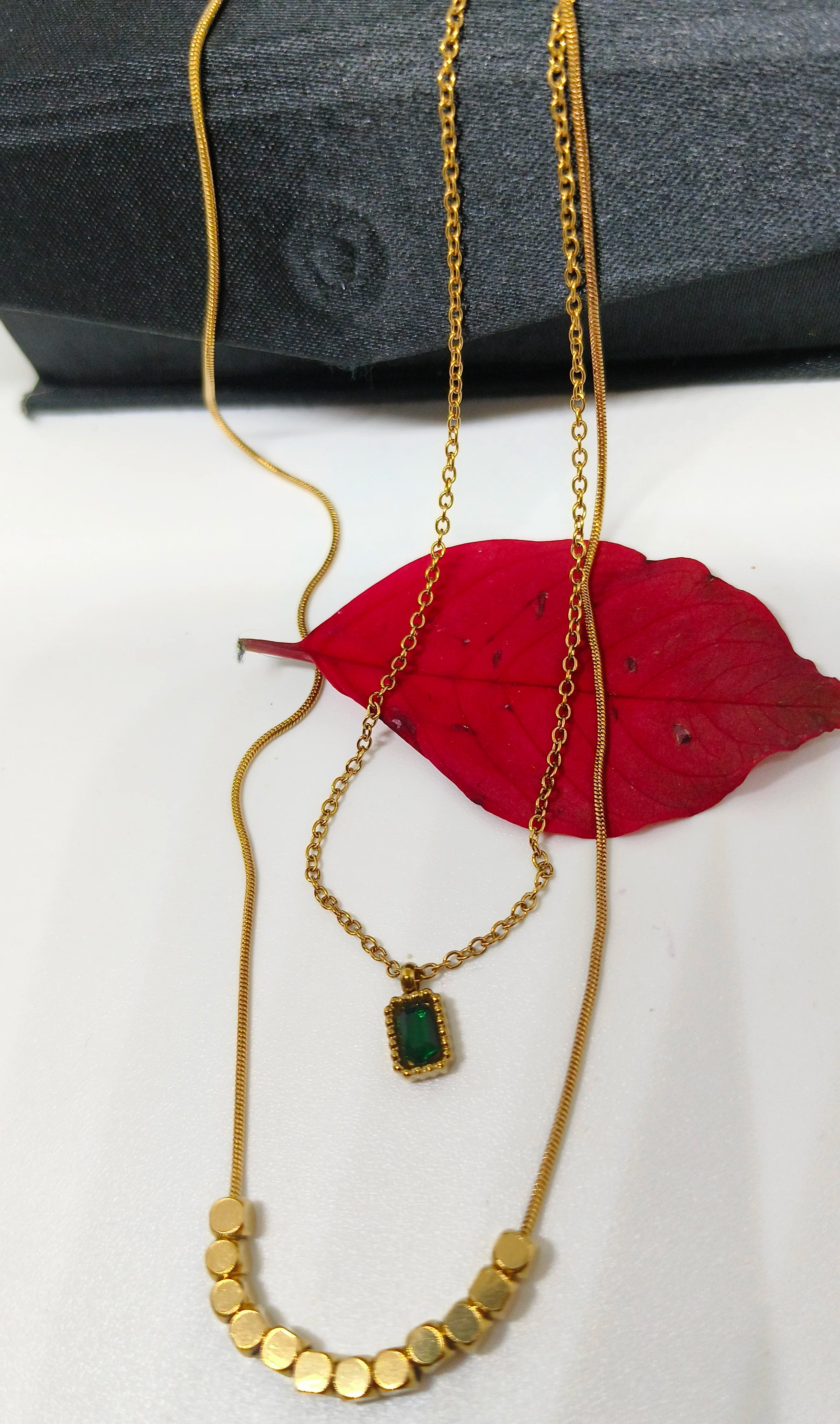 Green Stone Layered Necklace featuring a delicate gold chain with a small rectangular green stone pendant and a second gold chain with circular gold accents, displayed on a white surface with a red leaf and a black fabric background.