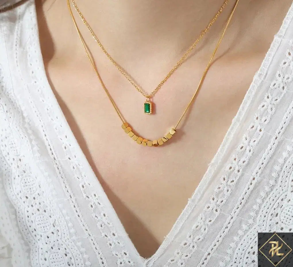 Close-up of a person wearing the Green Stone Layered Necklace featuring a gold chain with a small green rectangular stone pendant and a second gold chain with small gold beads, styled with a white textured blouse.
