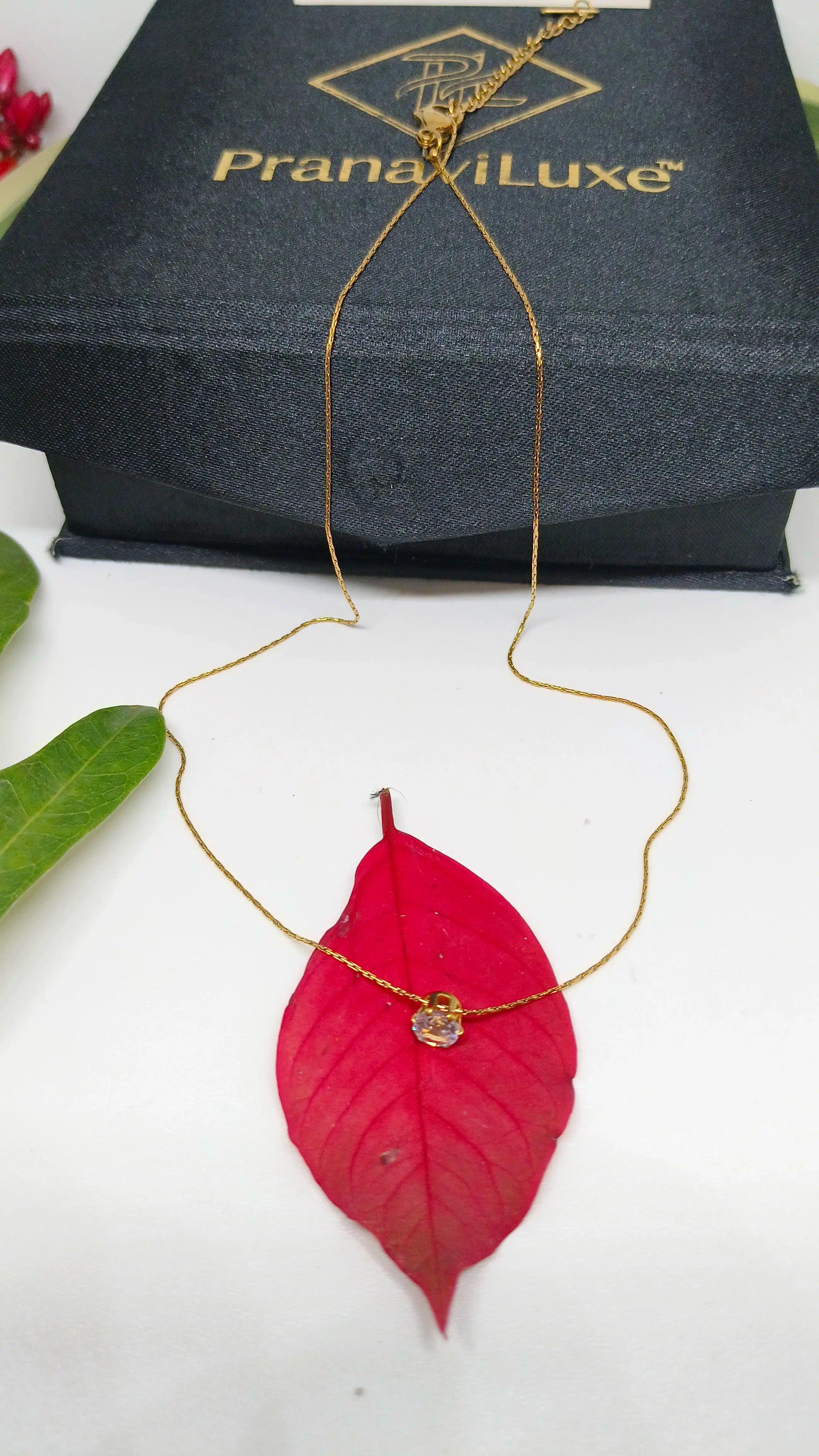 Gold necklace with a red leaf pendant on a white background, displayed in front of a PranaviLuxe box.