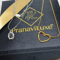 Two delicate necklaces displayed on a black branded box with a gold ribbon. One necklace is silver-toned with a faceted dark oval stone pendant and a small pearl bead.