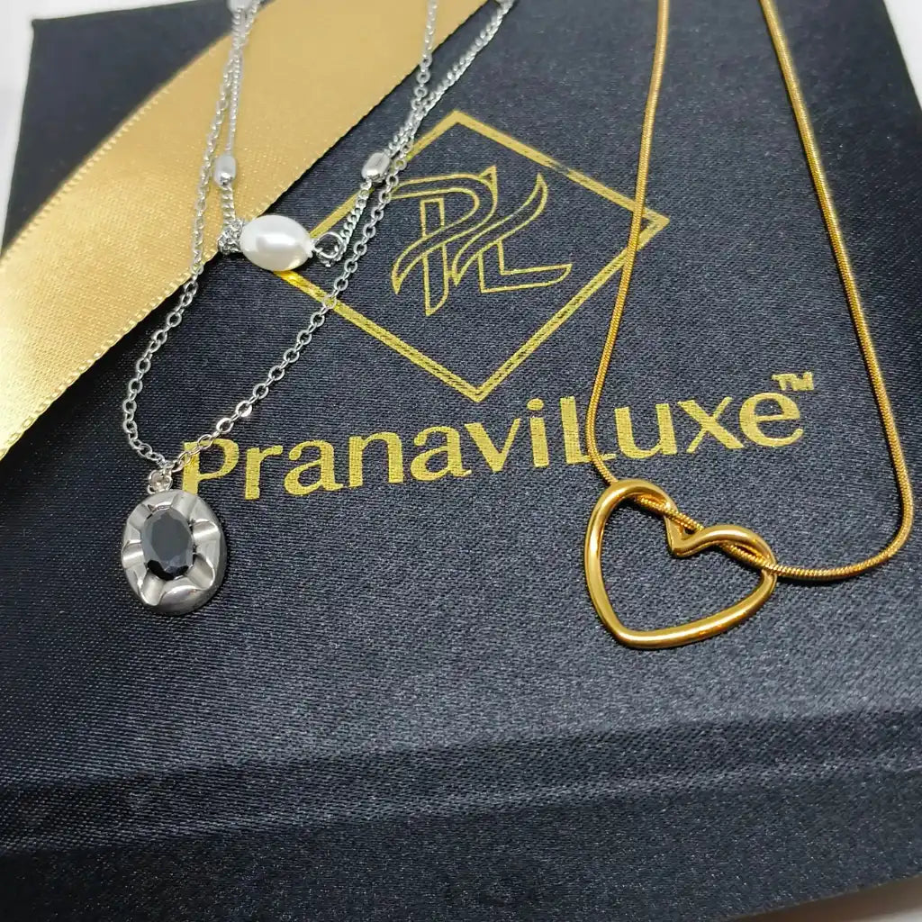 Two delicate necklaces displayed on a black branded box with a gold ribbon. One necklace is silver-toned with a faceted dark oval stone pendant and a small pearl bead.