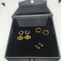 A set of gold-toned earrings displayed inside an open black velvet jewelry box. The set includes three pairs of hoop and dangle earrings, with two pairs featuring evil eye motifs with small blue and white details.