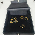 A set of gold-toned earrings displayed inside an open black velvet jewelry box. The set includes three pairs of hoop and dangle earrings, with two pairs featuring evil eye motifs with small blue and white details.