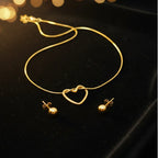 Gold necklace and matching stud earrings displayed on a black velvet surface with warm blurred bokeh lights in the background. The necklace features an open heart pendant, and the earrings have small round gold beads.