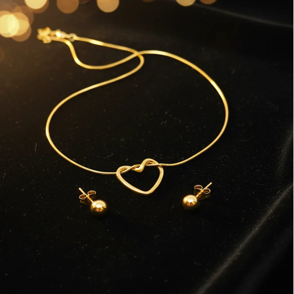 Gold necklace and matching stud earrings displayed on a black velvet surface with warm blurred bokeh lights in the background. The necklace features an open heart pendant, and the earrings have small round gold beads.