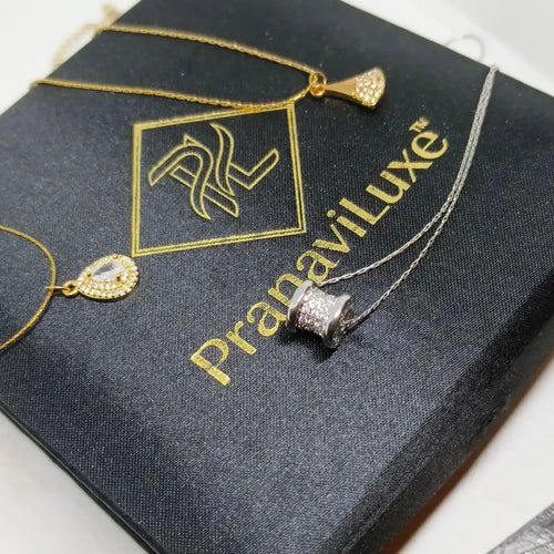Gold and silver necklace jewelry displayed on a black branded box with the text 'Pranavi Luxe'. The jewelry includes a gold chain with a small gold heart-shaped pendant, a gold chain with a gold pendant, and a silver chain with a silver cylindrical pendant embellished with clear stones.