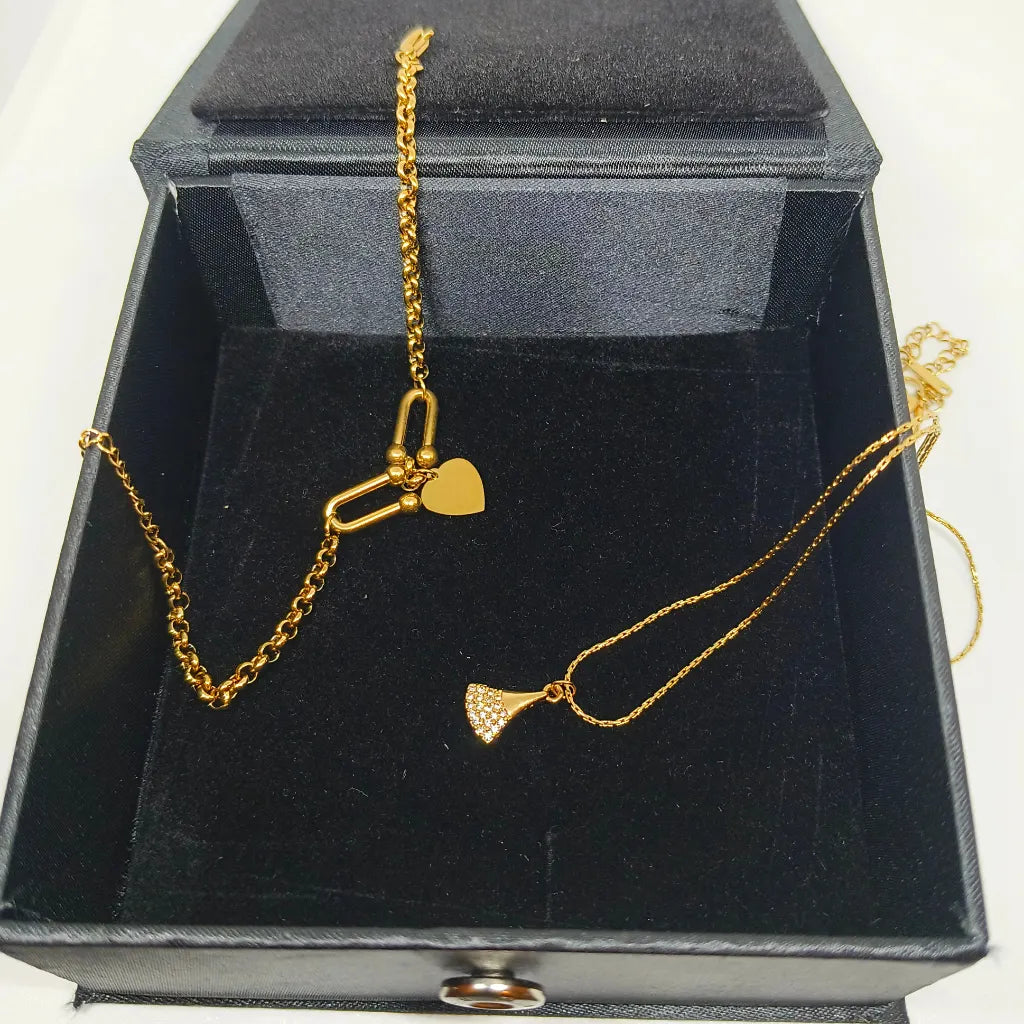 A gold necklace with two charms, one a heart and one a textured fan, displayed inside an open black velvet jewelry box.
