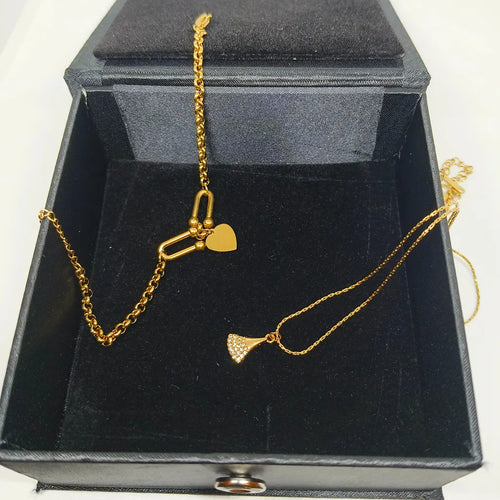 A gold necklace with two charms, one a heart and one a textured fan, displayed inside an open black velvet jewelry box.