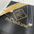 Two silver-toned necklaces displayed on a black jewelry box with gold branding. One necklace has a white pearl, one has a textured oval pendant with a dark center, and one has a cylindrical cluster pendant.