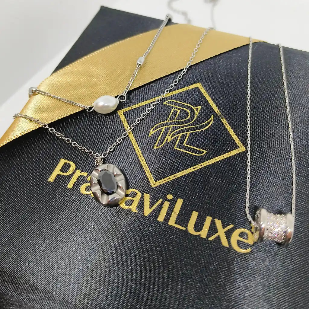 Two silver-toned necklaces displayed on a black jewelry box with gold branding. One necklace has a white pearl, one has a textured oval pendant with a dark center, and one has a cylindrical cluster pendant.