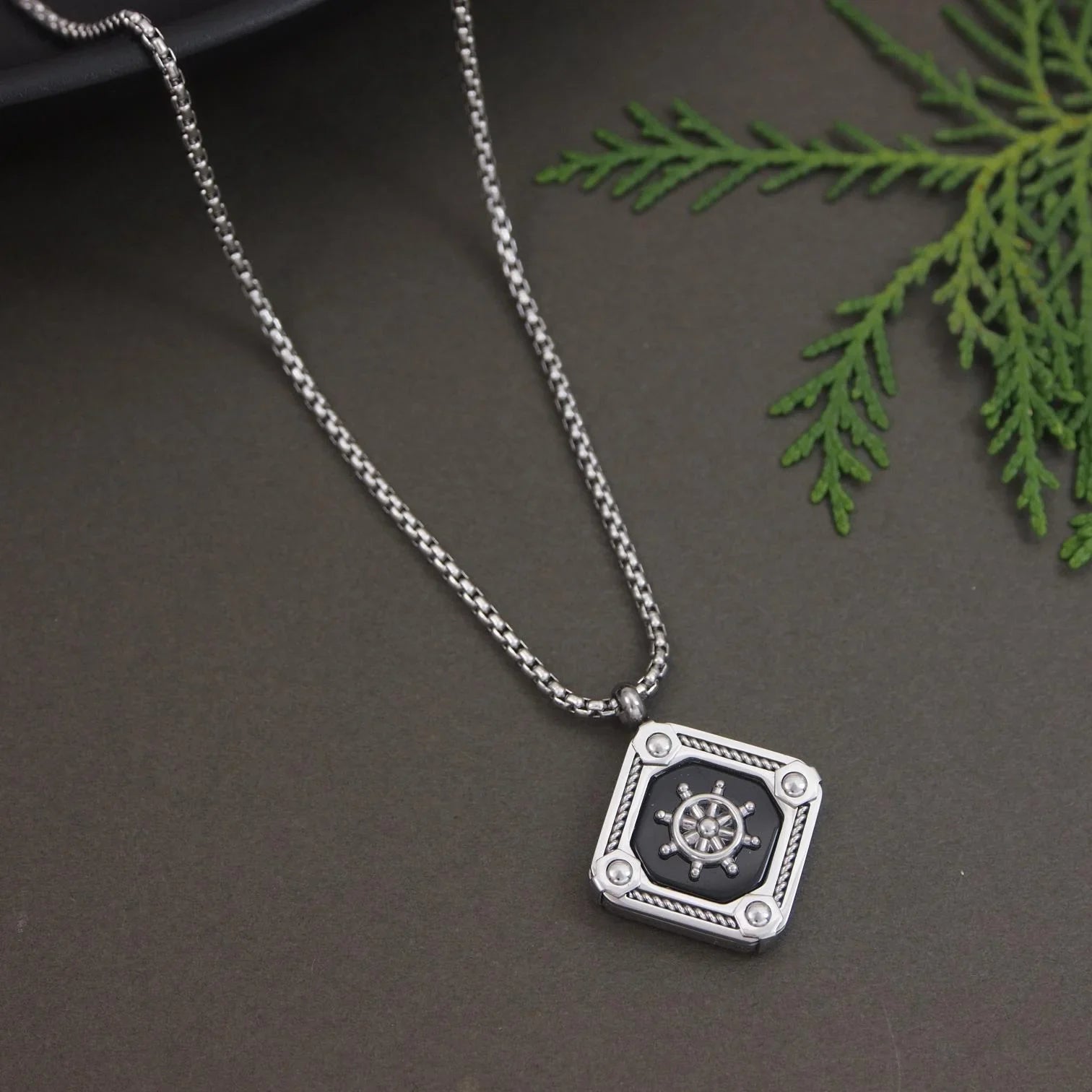 Silver necklace with a pendant featuring a ship's wheel design on a dark surface with green leaves.