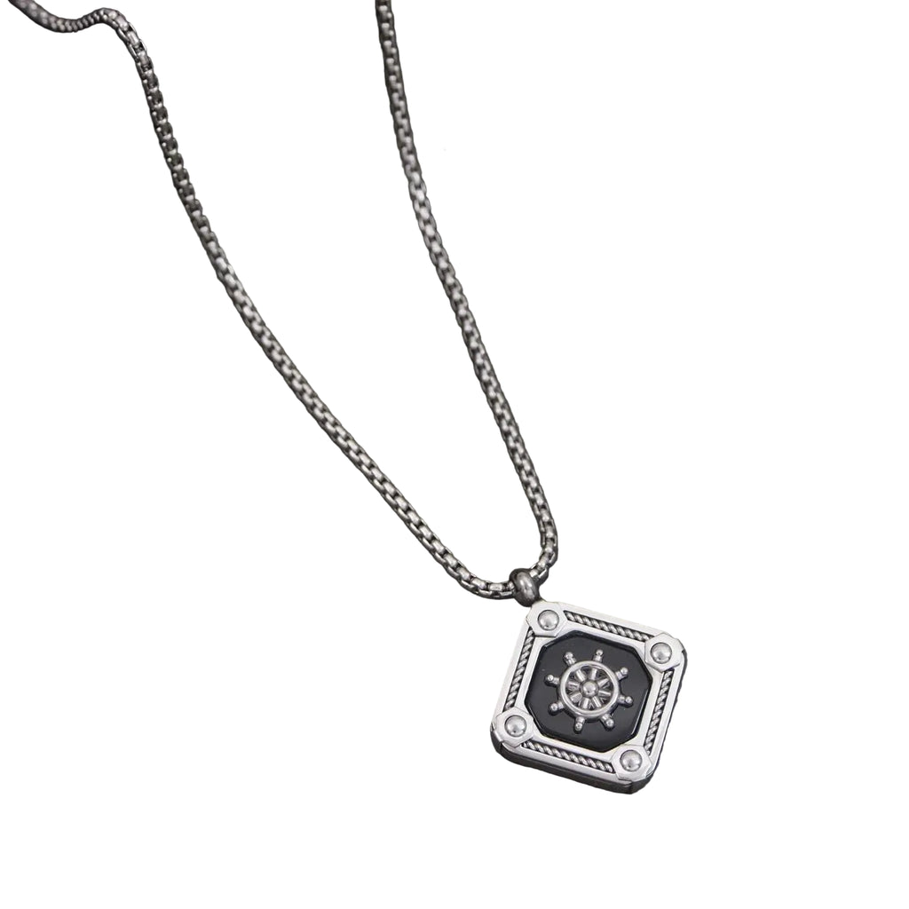 Silver necklace with a pendant featuring a ship's wheel design on a dark surface with green leaves.