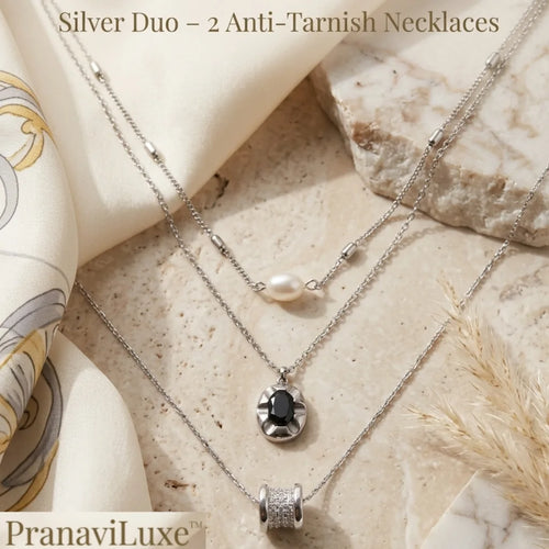 Two silver-toned anti-tarnish necklaces displayed on a beige stone surface with a cream silk scarf and dried grass. One necklace has a small white pearl, and the other has a round black stone pendant and a cylindrical pavé-set silver bead.