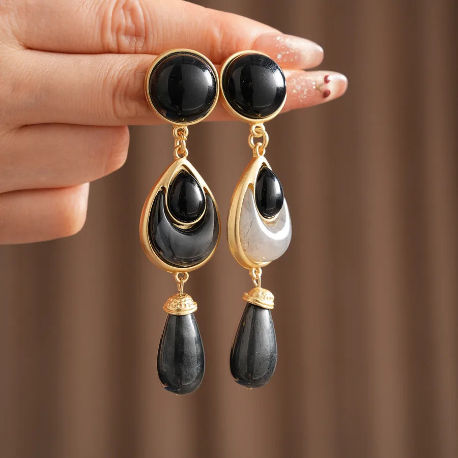 Black teardrop earrings with gold accents held by a hand against a brown background