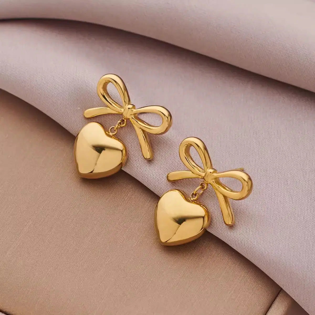 The Season of Bow Earrings stainless steel featuring elegant gold bow designs with dangling heart-shaped charms, perfect for stylish fashion jewelry and premium accessories.
