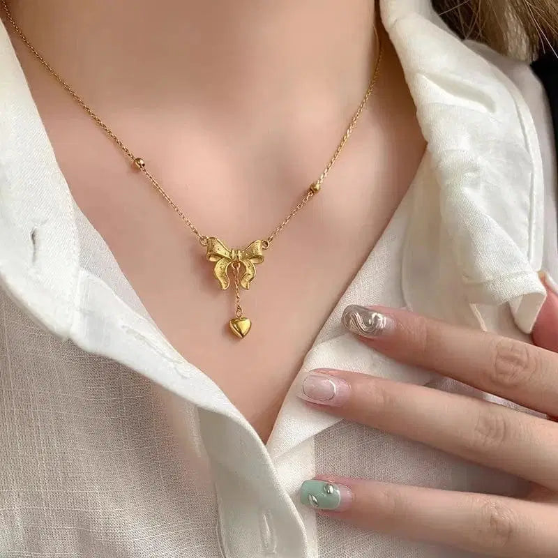 Gold necklace with a bow and heart pendant worn by a person in a white shirt.