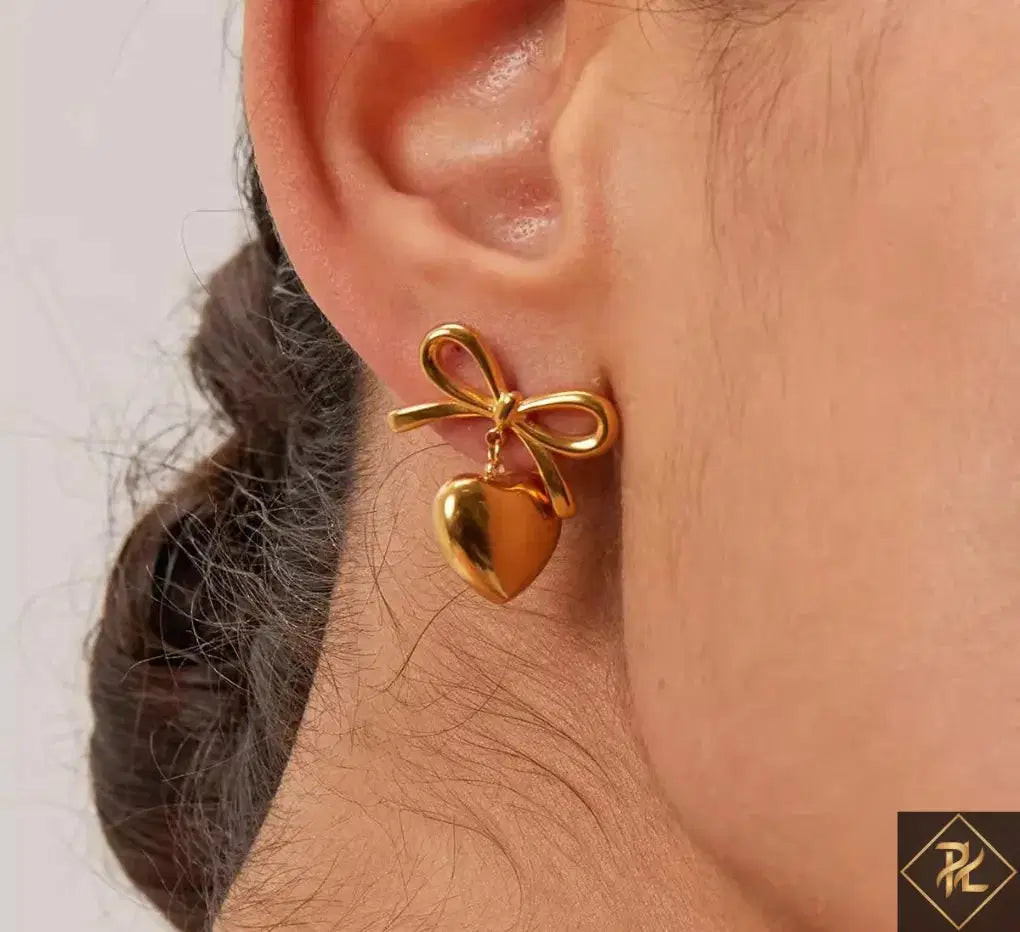 Close-up of an ear wearing The Season of Bow Earrings stainless steel, featuring a gold-toned bow with a dangling heart charm, stylish and elegant fashion jewelry.