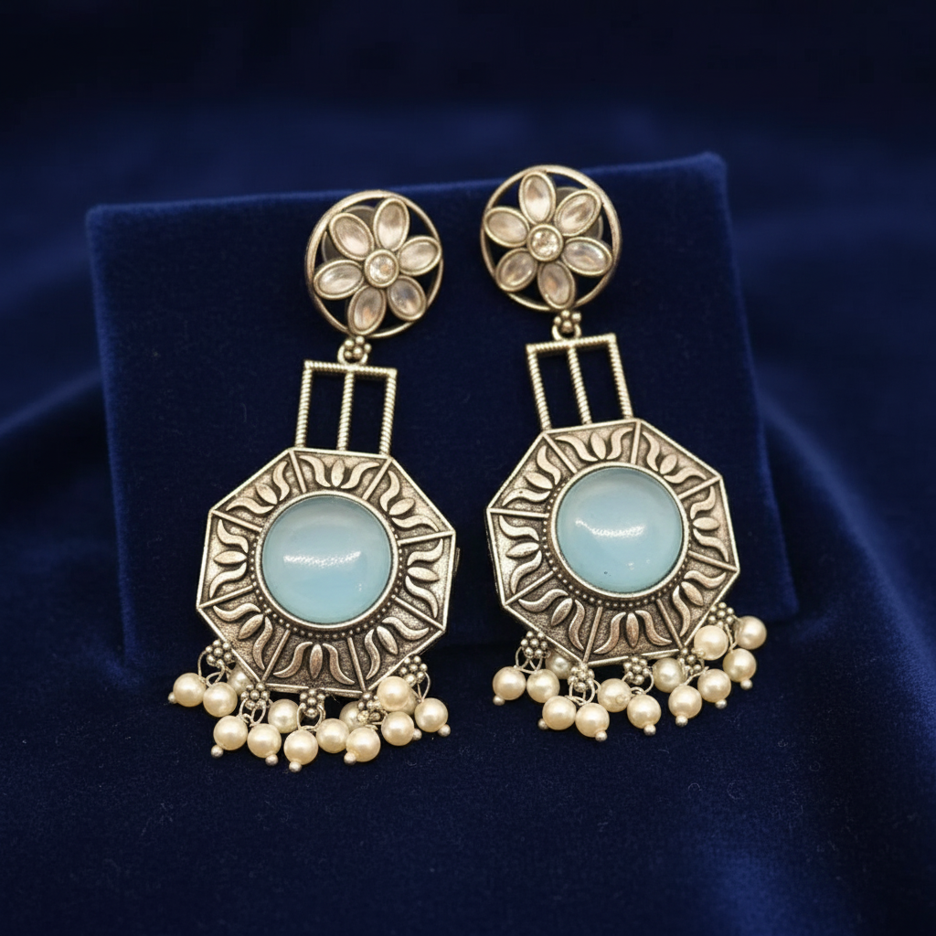 A pair of ornate gold-toned statement earrings with light blue oval stones and dangling pearl-like beads, displayed on a dark blue velvet stand.