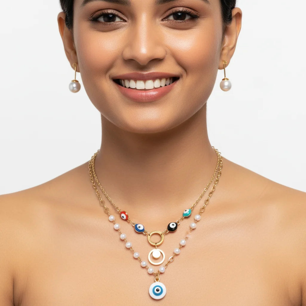A close-up of a woman wearing a gold and pearl double-layered necklace with colorful evil eye beads and a blue-and-white pendant, along with matching pearl drop earrings, against a light gray background.