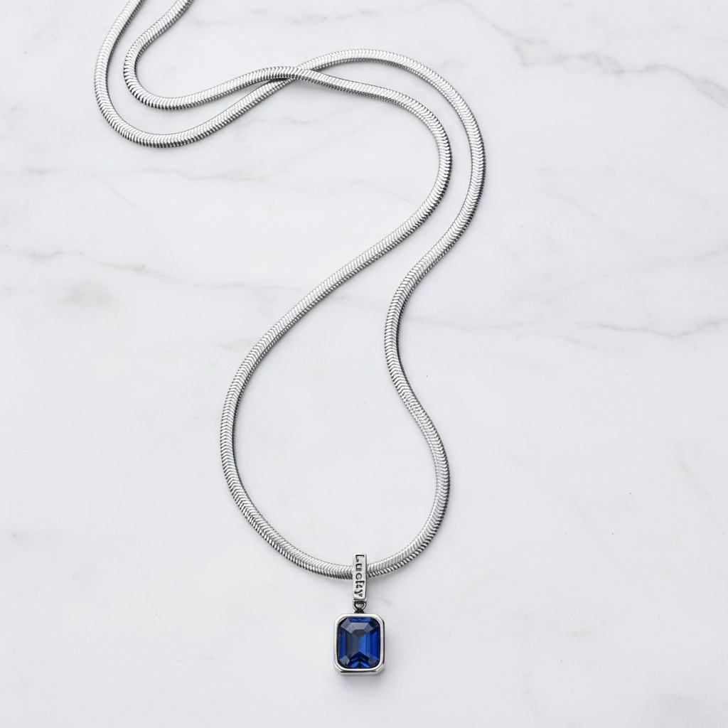 A silver snake chain necklace with a rectangular blue gemstone pendant, displayed on a white marble background.