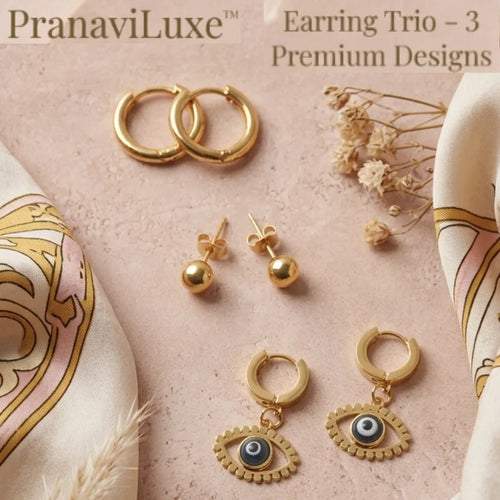 Three gold-tone earrings displayed on a soft pink textured background with decorative fabric and dried flowers. The set includes two hoop earrings, a pair of stud earrings with round beads, and a pair of hoop earrings with dangling evil eye charms.