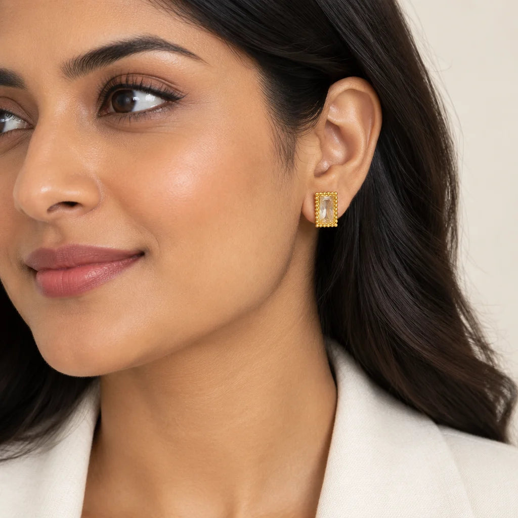 A close-up of a woman wearing a small gold rectangular stud earring with a clear central stone, shown against a soft beige background.
