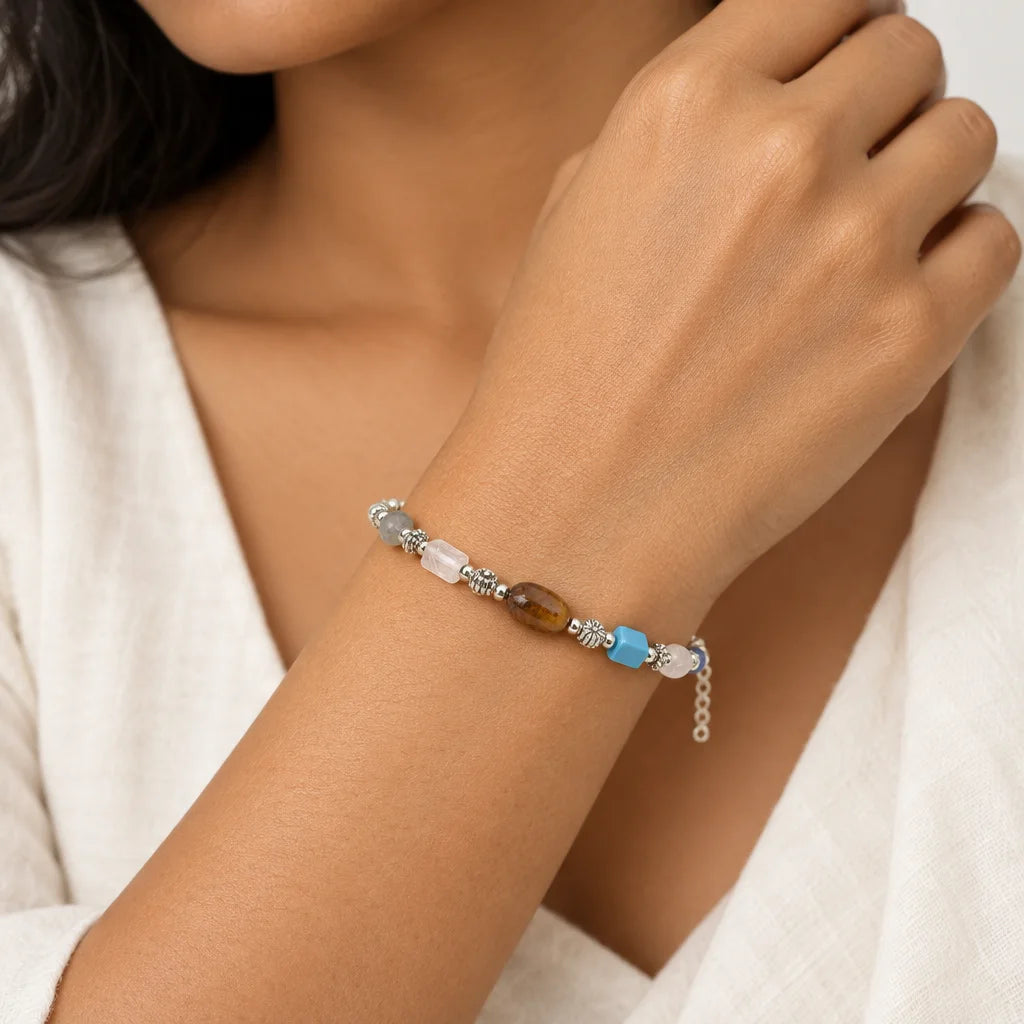 A close-up of a person's wrist wearing a delicate beaded bracelet. The bracelet features silver-toned metal links, clear faceted beads, a translucent white bead, a brown faceted bead, and two small light blue beads.