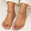 A close-up of a person's feet wearing black cord anklets decorated with small silver flower charms, styled with light beige linen pants.