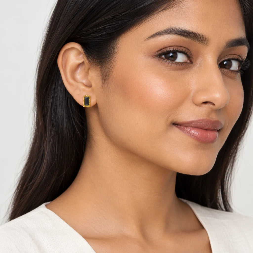 A close-up of a woman wearing a small rectangular gold earring with a dark green stone on her earlobe, set against a clean light gray background.