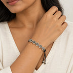 A close-up of a woman wearing a delicate silver flower bracelet with a black cord and bead tassel, paired with a white top.