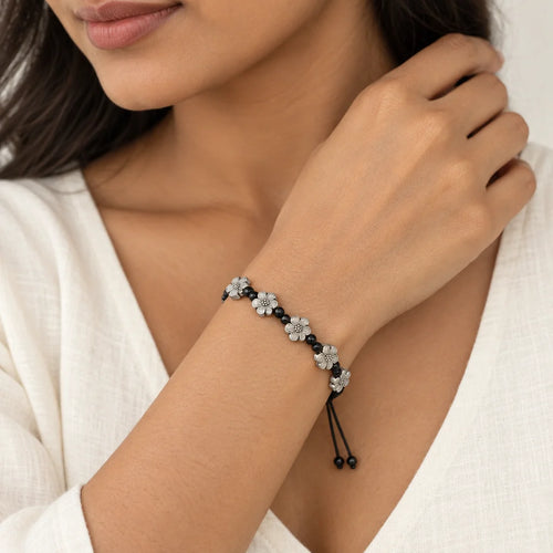 A close-up of a woman wearing a black cord bracelet with silver and white flower charms, styled with a white top.