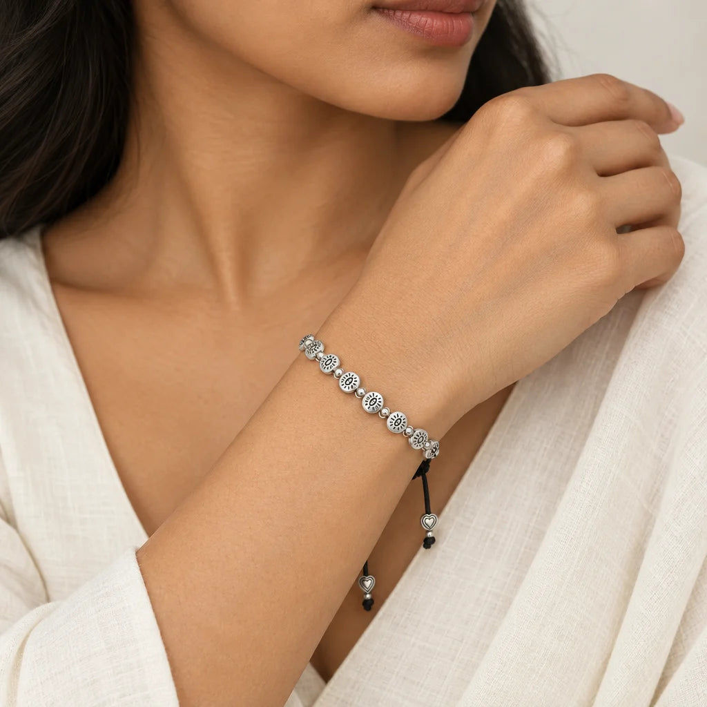 A close-up of a woman wearing a delicate silver beaded bracelet with clear stones and a black cord tassel, paired with a white linen top.
