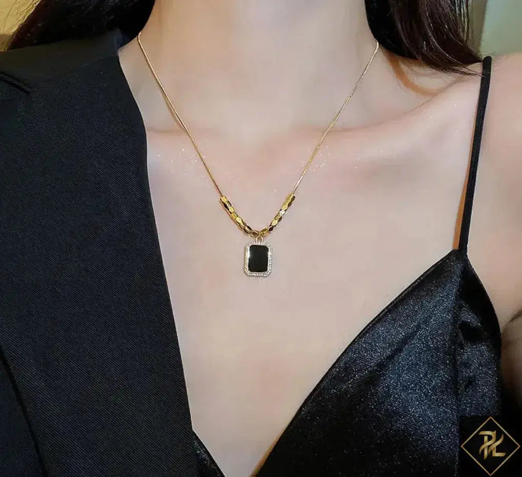 Gold necklace with a black pendant worn by a person in a black outfit.