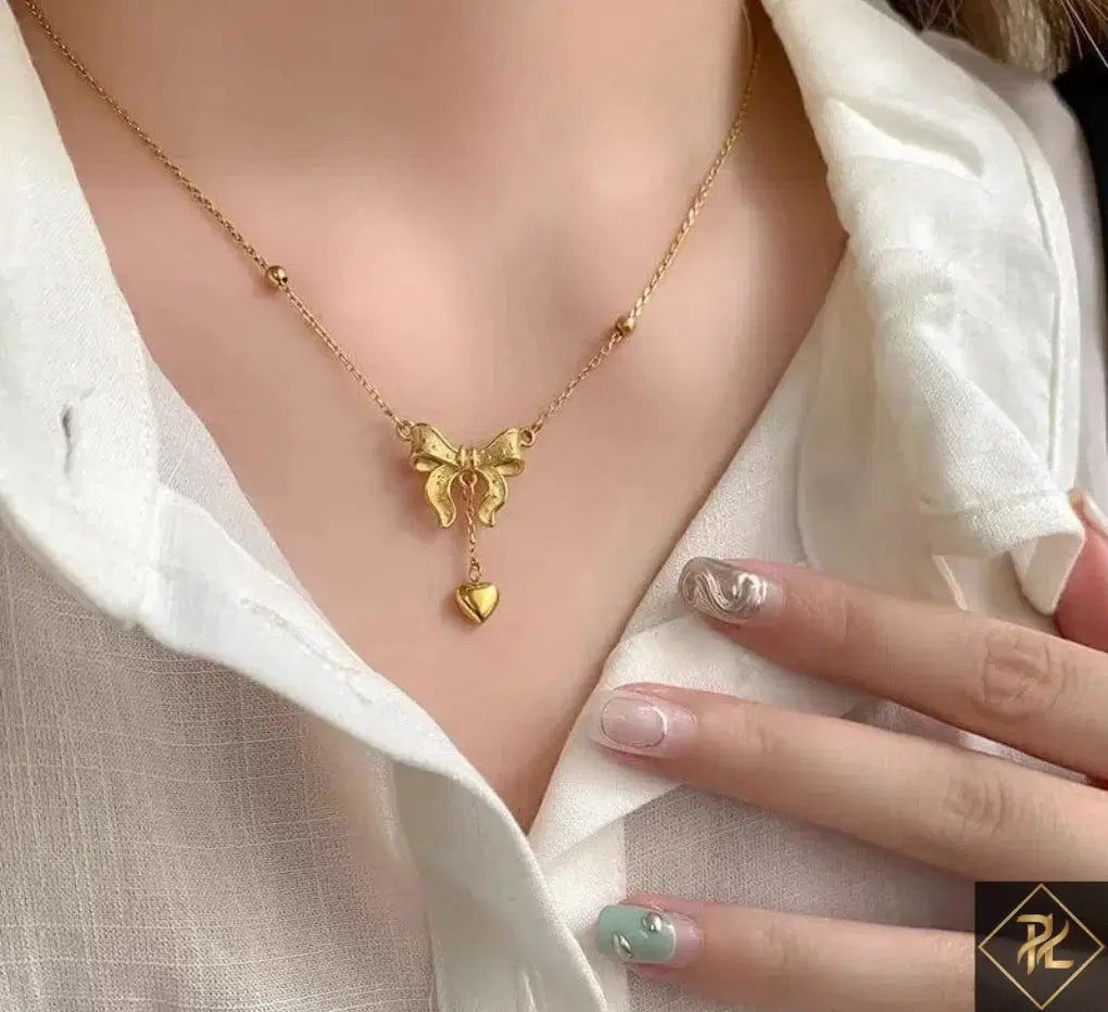Gold necklace with heart and bow design worn by a person in a white shirt.