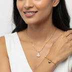 A woman wearing a delicate gold chain necklace with a small fan-shaped pendant and a matching gold bracelet with a small heart charm, styled with a white sleeveless top against a light gray background.