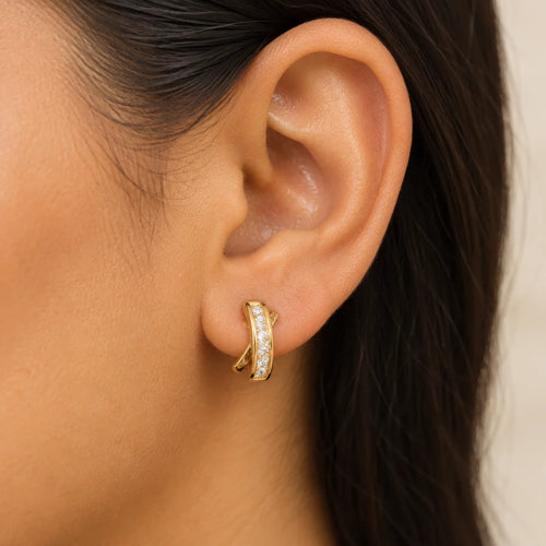 A close-up of a person's ear wearing a small gold huggie hoop earring set with clear stones, against a soft light beige background.