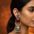 A close-up of a woman's ear wearing an ornate silver-toned jhumka earring with a round reddish-brown stone and dangling silver bells, against a warm brown background.