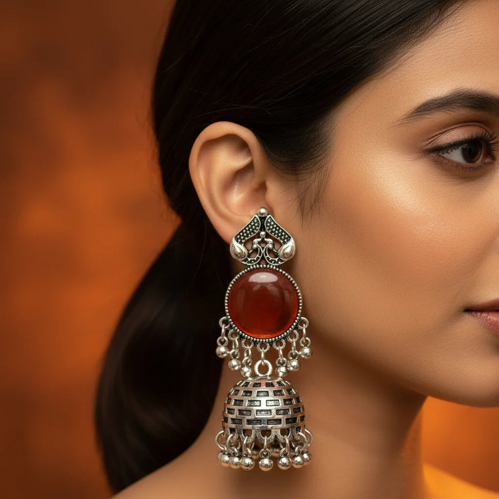 A close-up of a woman's ear wearing an ornate silver-toned jhumka earring with a round reddish-brown stone and dangling silver bells, against a warm brown background.